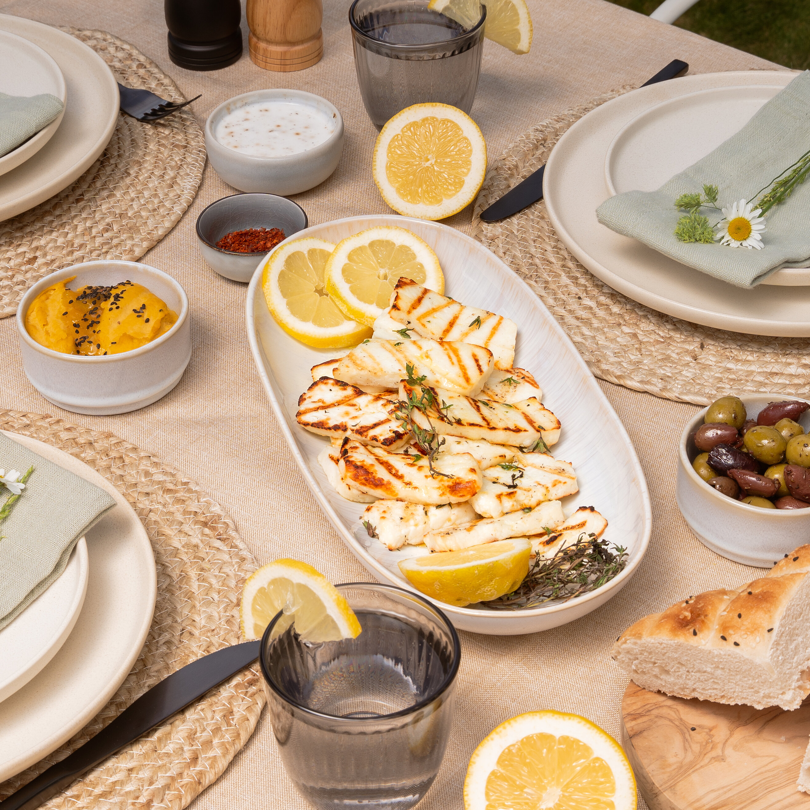 Set table with grilled halloumi, lemon slices, bread, olives, and fresh flowers.
