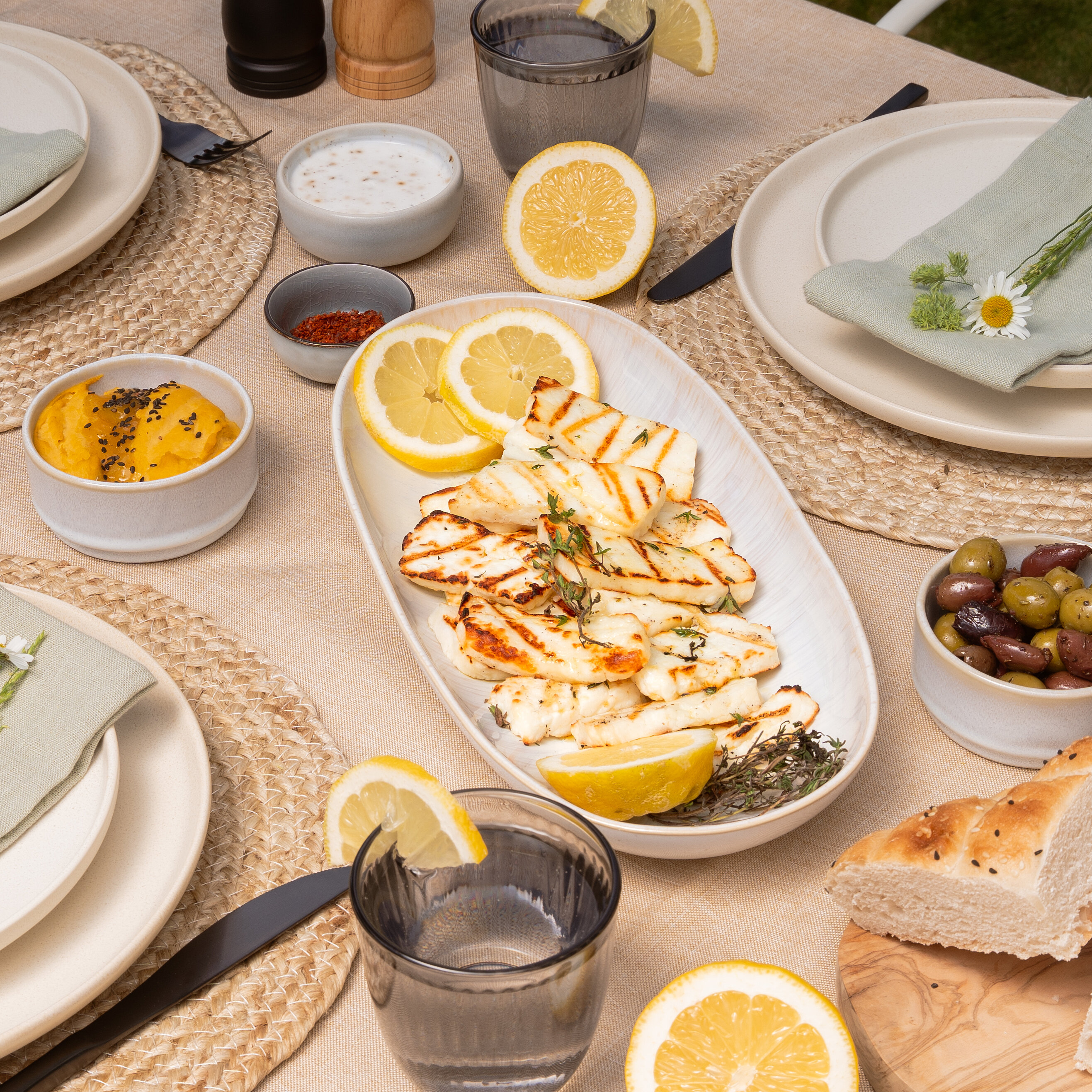 Gedeckter Tisch mit gegrilltem Halloumi, Zitronenscheiben, Brot, Oliven und frischen Blumen.