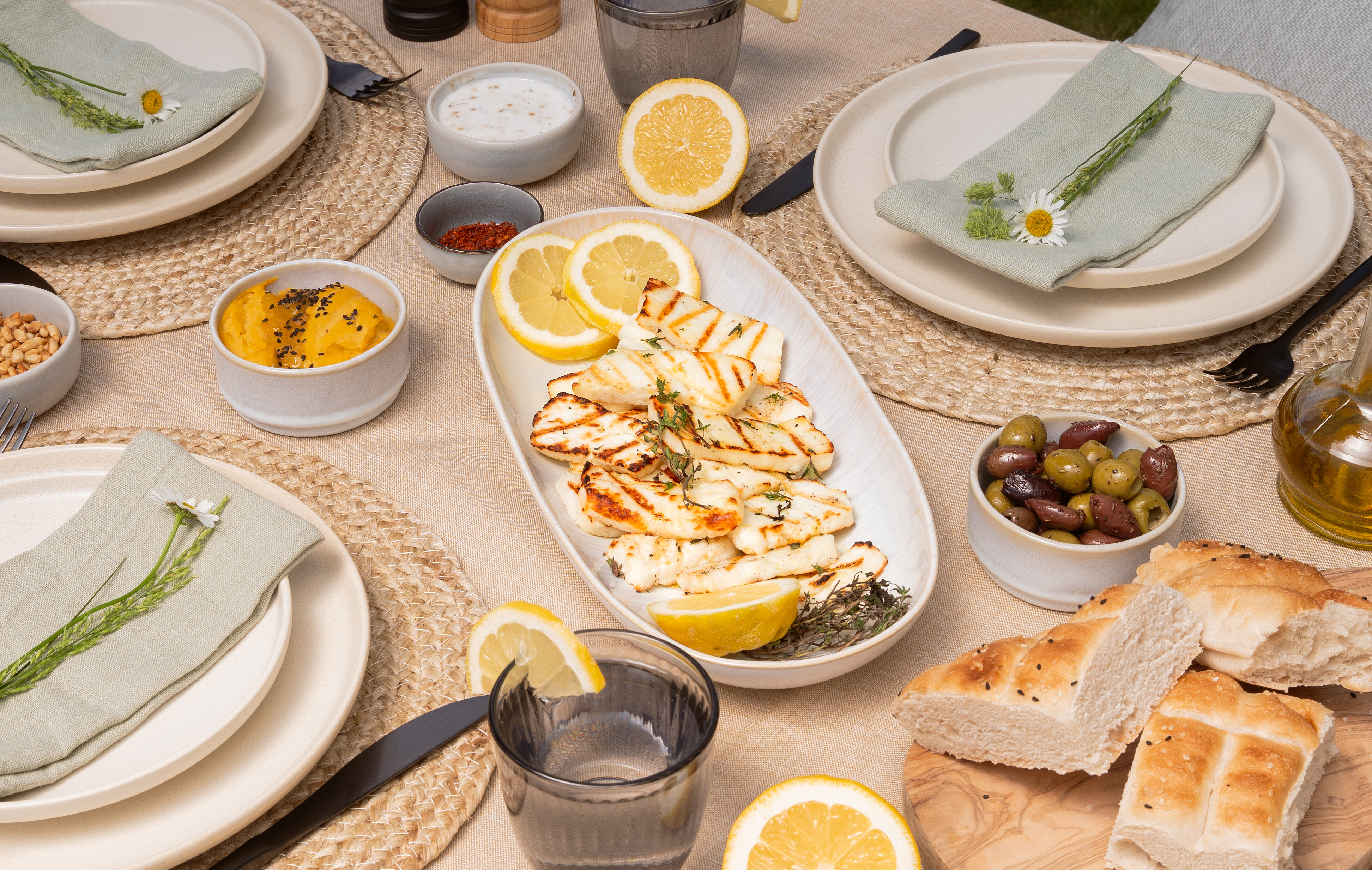 Set table with grilled halloumi, lemon slices, bread, olives, and fresh flowers.